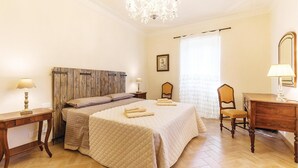 Une chambre d’hôtel avec un grand lit, deux tables de chevet, une tête de lit en bois, un lustre, un bureau avec une chaise, un miroir et une fenêtre avec des rideaux légers.