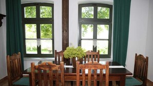 A dining area with a wooden table and chairs, two windows with curtains, and a potted plant on the table. - Apartment IV - Apartments in farmhouse - Property 44365 (Papendorf)