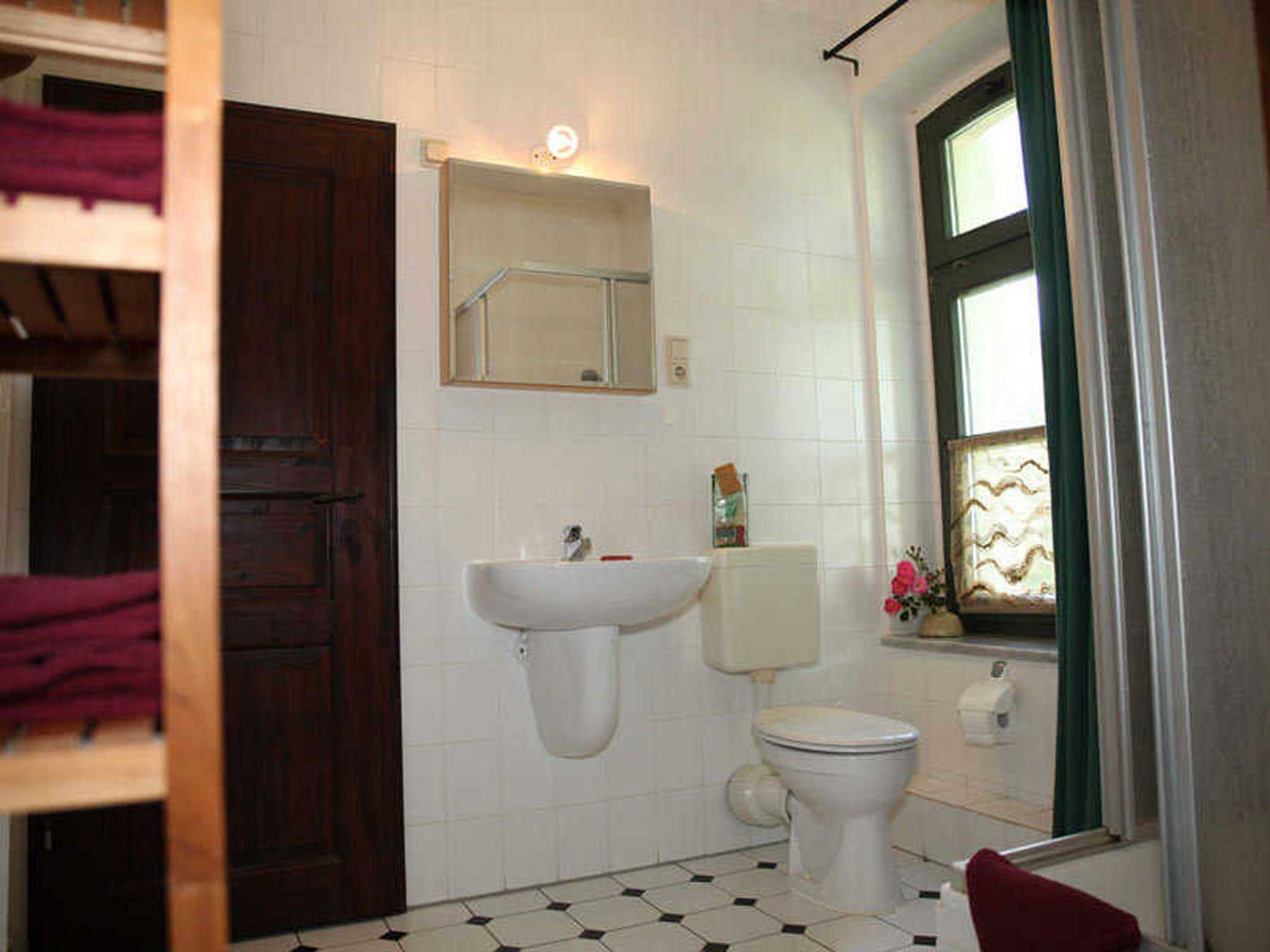 A bathroom with a white sink, toilet, and a mirror above the sink. There is a window with a curtain and a towel rack with a red towel.