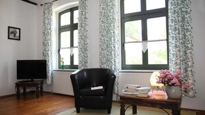 A living room with a black leather armchair, a wooden coffee table with books and a vase of flowers, a television on a stand, and large windows with patterned curtains. - Apartment IV - Apartments in farmhouse - Property 44365 (Papendorf)