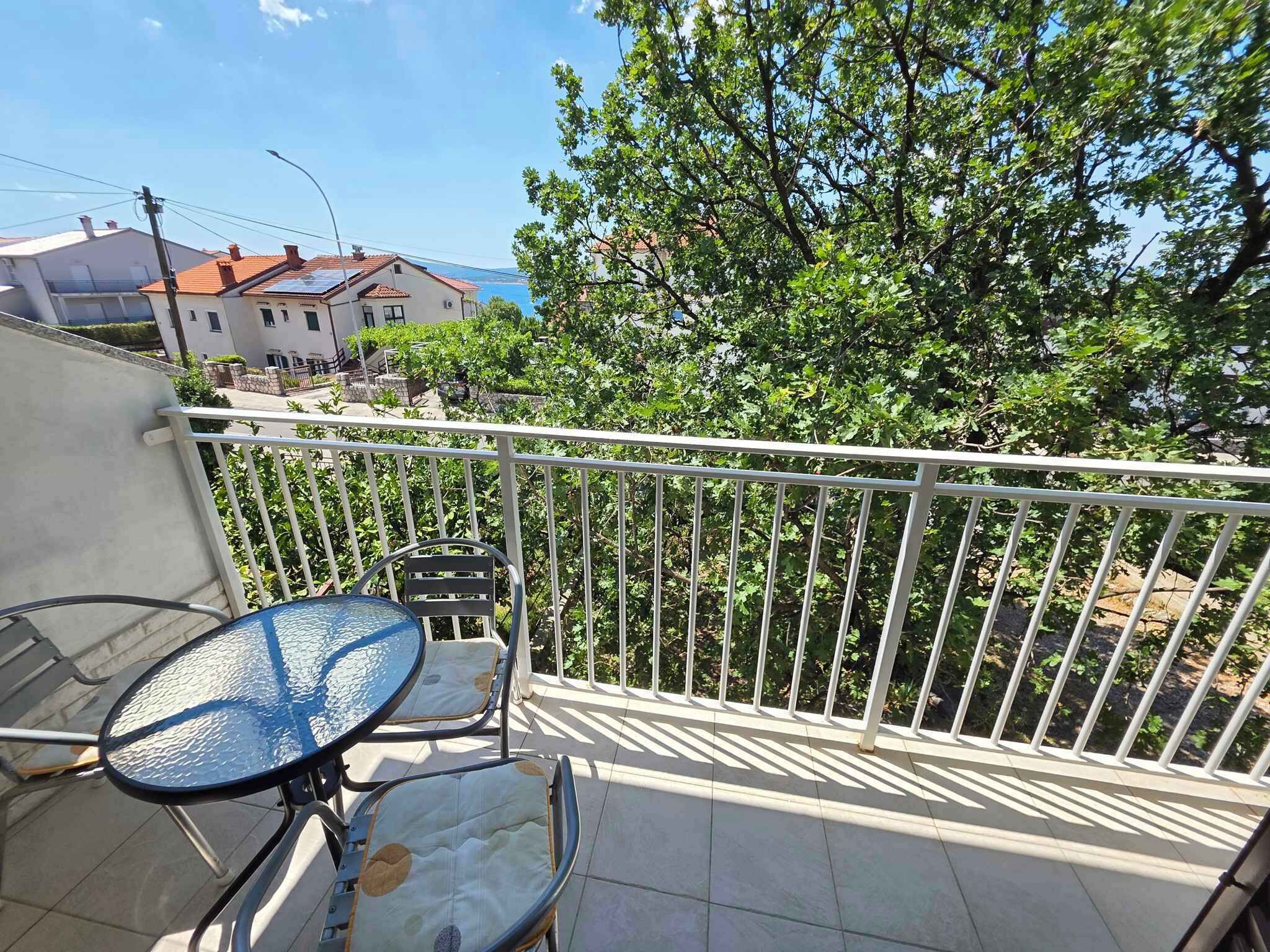 Ein Balkon mit einem Metalltisch und Stühlen, einem weißen Geländer und Blick auf Gebäude und Bäume.