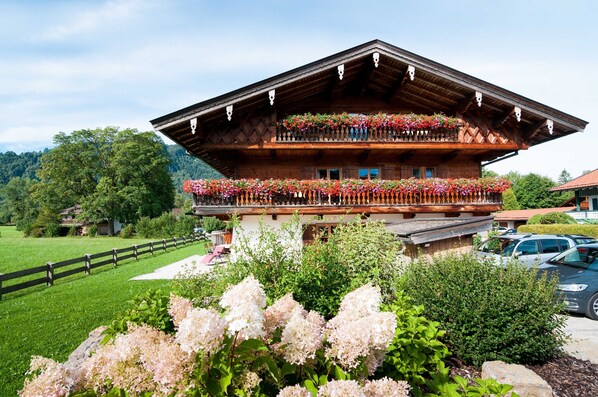 A traditional wooden chalet with a balcony adorned with flowers, surrounded by lush greenery and a clear sky.