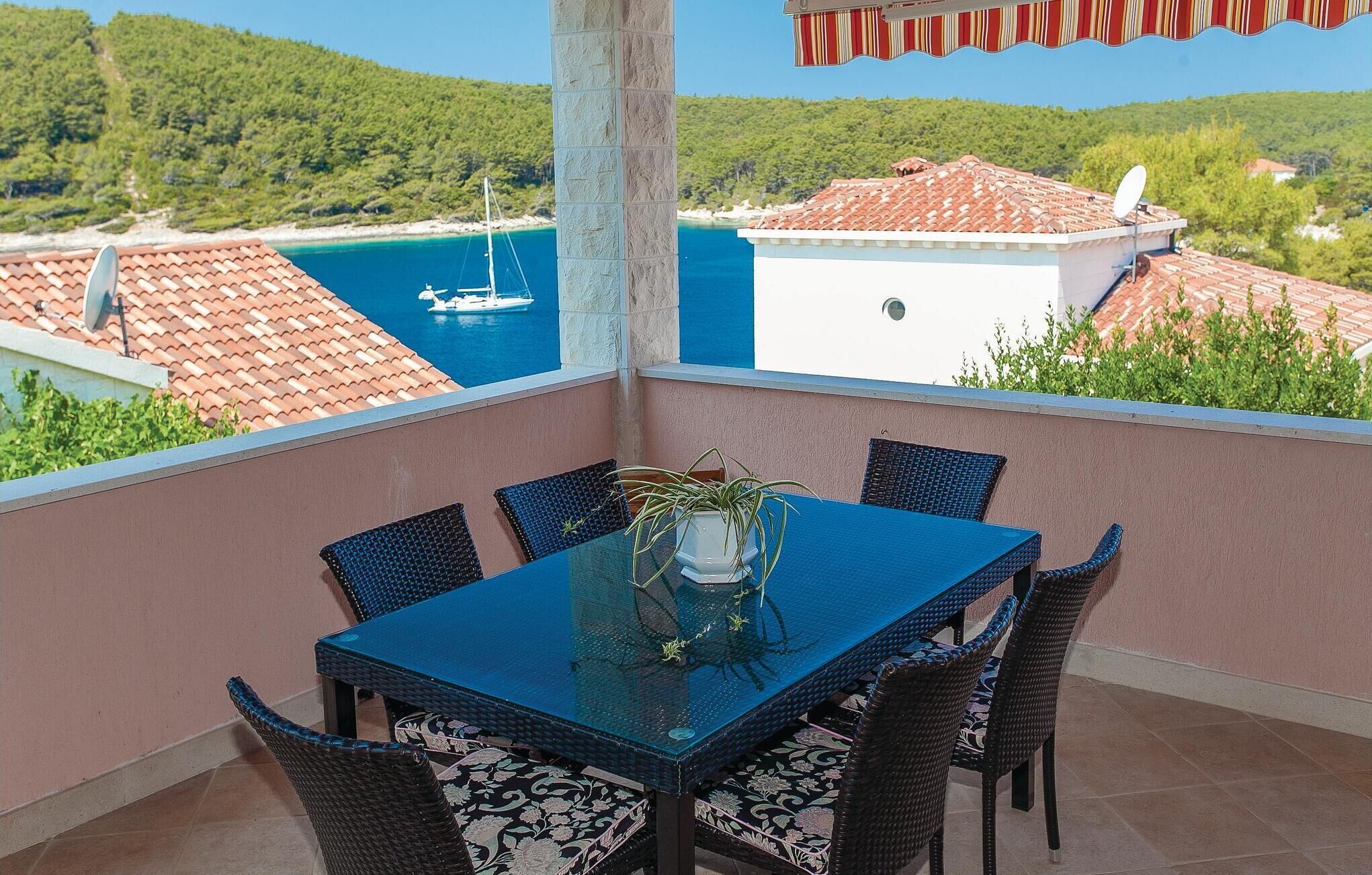 A balcony with a glass table and wicker chairs, overlooking a coastal area with a boat and buildings.