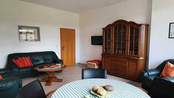 Ein Wohnzimmer mit einem schwarzen Ledersofa, einem Holzkabinett, einem runden Tisch mit kariertem Tischläufer und einem Deckenventilator.
