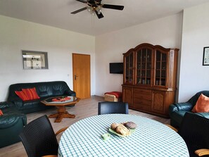 Ein Wohnzimmer mit einem schwarzen Ledersofa, einem Holzkabinett, einem runden Tisch mit kariertem Tischläufer und einem Deckenventilator.