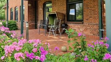 Ein Backsteinhaus mit überdachter Terrasse, Außenbestuhlung und blühenden Pflanzen.