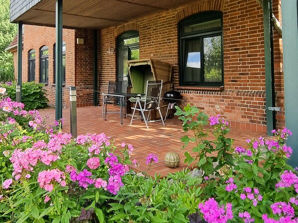 Edificio de ladrillo con un porche cubierto, asientos al aire libre y plantas con flores.