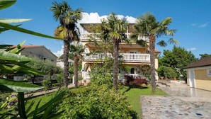 A multi-story building with a balcony, surrounded by palm trees and a well-maintained garden. - Holiday apartment with shared swimming pool (Malinska)