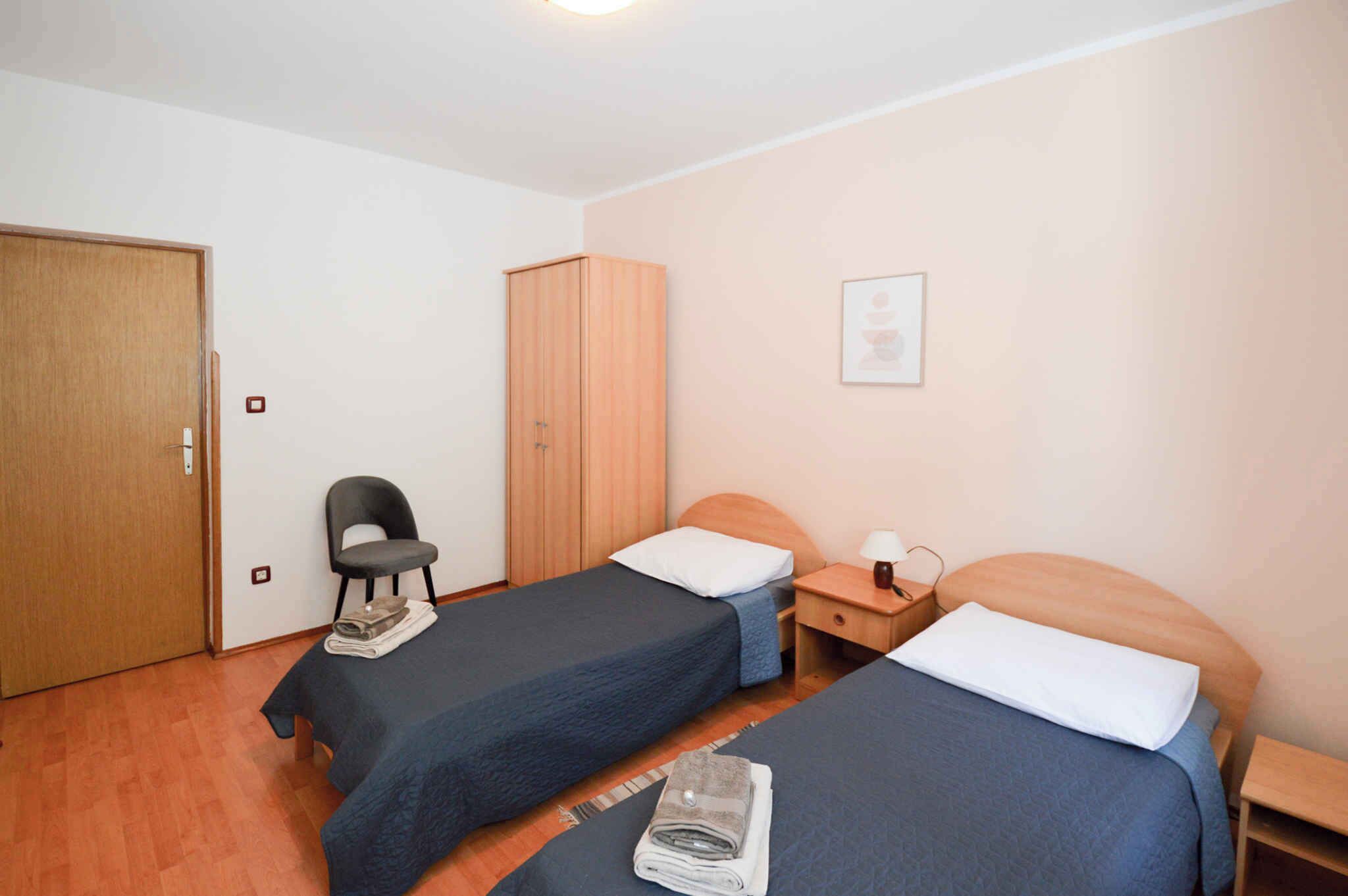 A room with two single beds, a wooden wardrobe, a chair, a bedside table with a lamp, and a framed picture on the wall.