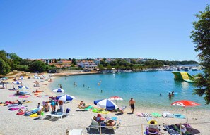 A beach with people sunbathing and swimming, umbrellas, and boats in the background.