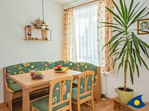 Ein gemütlicher Essbereich mit einem Holztisch, grünen Polsterstühlen und einer Obstschale. Außerdem ist ein Blumentopf und ein Regal mit Dekorationsgegenständen zu sehen.