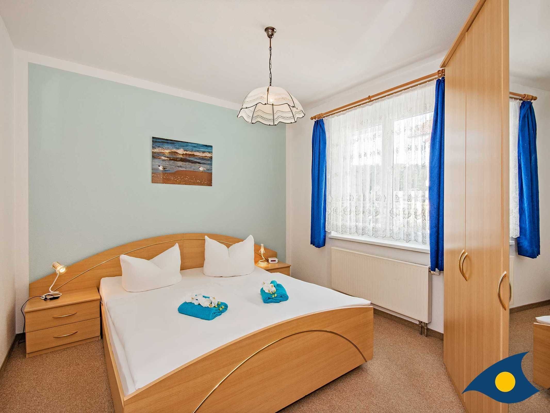 Une chambre avec un lit en bois, une table de chevet, une commode, une fenêtre avec des rideaux bleus et un tableau représentant une plage accroché au mur.