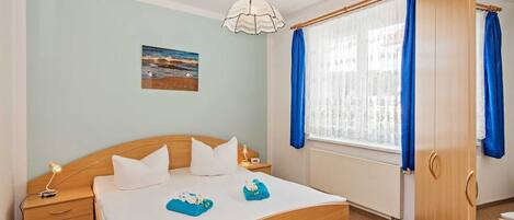 Une chambre avec un lit en bois, une table de chevet, une commode, une fenêtre avec des rideaux bleus et un tableau représentant une plage accroché au mur.