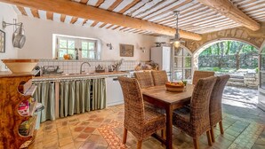 A rustic kitchen with wooden beams, a dining area with wicker chairs, and a view of the garden through an arched doorway.