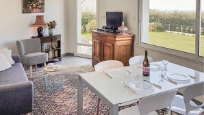 Un accogliente soggiorno con un divano, una poltrona a fantasia, un tavolino da caffè e un tavolo da pranzo per due, apparecchiato con bicchieri da vino e una bottiglia. Si notano anche una libreria e un mobile porta televisione.