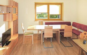 Ein Zimmer mit Holztisch, Stühlen, einer Couch, einem Fernseher und einem Fenster mit Blick ins Grüne.