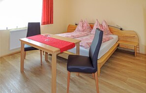 Ein Schlafzimmer mit einem hölzernen Bett, einem kleinen Tisch mit rotem Tischlein, zwei Stühlen und einem Fenster mit roten Vorhängen.
