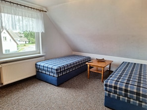 A small, triangular room with a window, a radiator, a bed with a plaid cover, a small wooden table, and a bench.