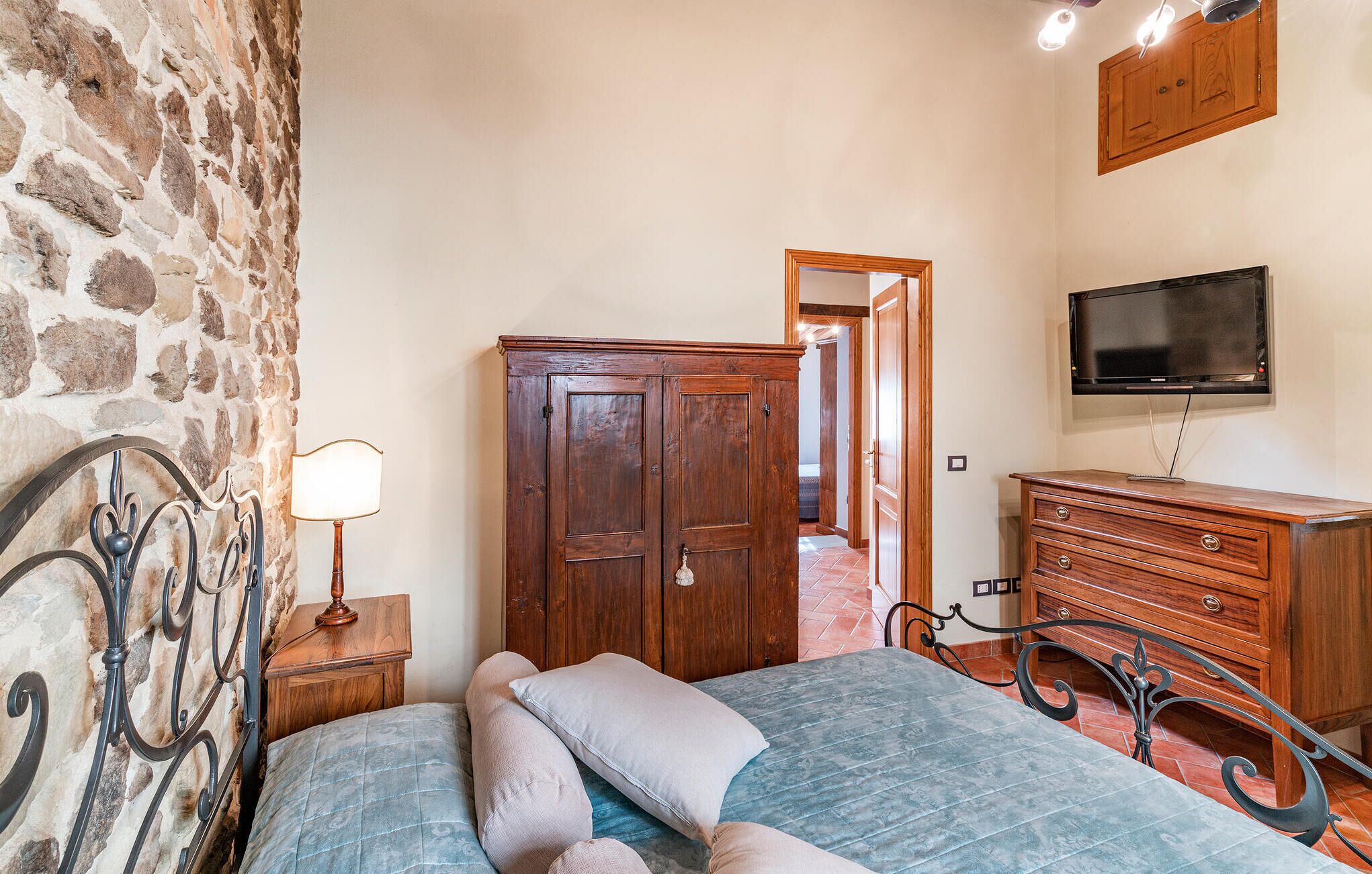 A bedroom with a stone wall, a bed with pillows, a wooden wardrobe, a TV, and a nightstand with a lamp.