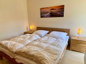 Una cama bien hecha con sábanas blancas, dos mesitas de noche con lámparas y un cuadro de un atardecer en la pared.