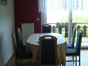 Ein Essbereich mit einem runden Tisch, Stühlen, einer weißen Tischdecke und Blick auf einen Balkon mit Geländer.