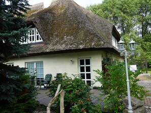 A traditional thatched-roof house with a white exterior, a wooden deck, and a garden with plants and a bench.