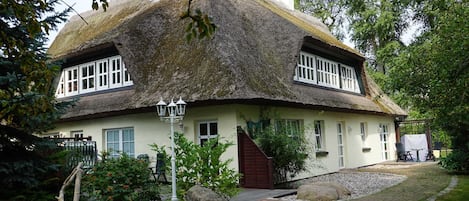 Une maison traditionnelle à toit de chaume, avec une façade blanche, un escalier en bois menant à l’entrée et un chemin pavé devant.