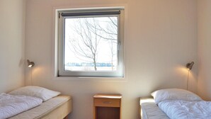 A bedroom with two single beds, a small wooden nightstand, a window with a view of bare trees, and wall-mounted reading lamps. - 2 bedroom cozy home in Rømø (Rømø)