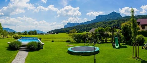 A well-maintained lawn with a swimming pool, a trampoline, and a small house with a red roof.