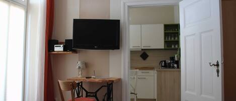 A hotel room with a bed, a desk with a chair, a television, and a kitchenette in the background.