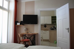 Quarto de hotel com uma cama, uma escrivaninha com cadeira, televisão e uma kitchenette ao fundo.
