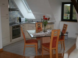 A compact kitchen with a dining area, featuring a glass table and wooden chairs, a vase with flowers, and a view through a window.
