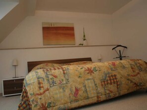 A bedroom with a bed covered in a floral-patterned blanket, a bedside table with a lamp, and a wall-mounted shelf with a plant. - Apartment Im Alten Gut (Erfurt)