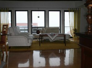 A living room with a white sofa, a wooden coffee table, and a chair. There are large windows with curtains, and a view of the outside. - Apartment Im Alten Gut (Erfurt)