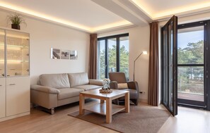 A modern living room with a sofa, a coffee table, a cabinet with glass doors, and large windows offering a view of trees. - Stunning apartment in Binz with house sea view (Prora/Rügen)
