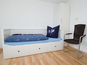 Une chambre avec un lit simple, une tête de lit blanche, un matelas bleu, un oreiller bleu foncé, une chaise noire et une armoire blanche.