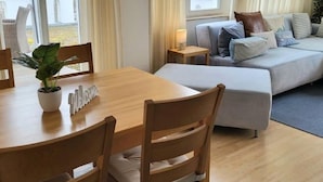 Une pièce comprenant une table à manger en bois, un canapé, une petite table de chevet avec une lampe et une fenêtre avec des rideaux.
