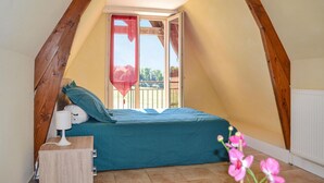 Une petite chambre mansardée de forme triangulaire, avec un lit, une table de chevet et une fenêtre donnant sur un balcon.