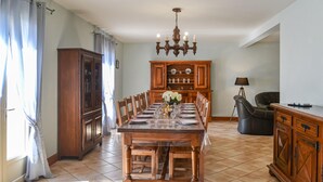 Un coin repas avec une table en bois dressée pour six personnes, entourée de chaises. Un lustre se trouve au-dessus de la table, et on aperçoit un buffet rempli de vaisselle au fond de la pièce.