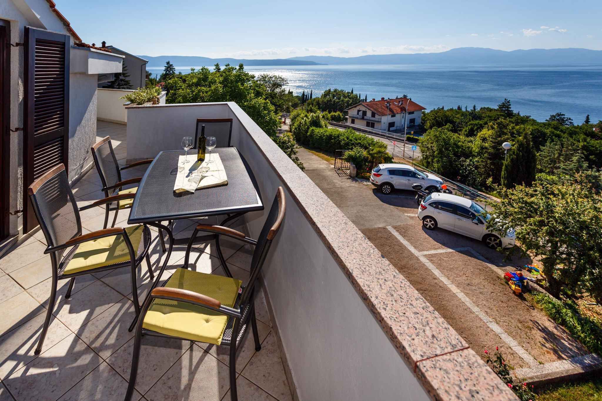 A balcony with a table and chairs, overlooking a parking area and a view of the sea.