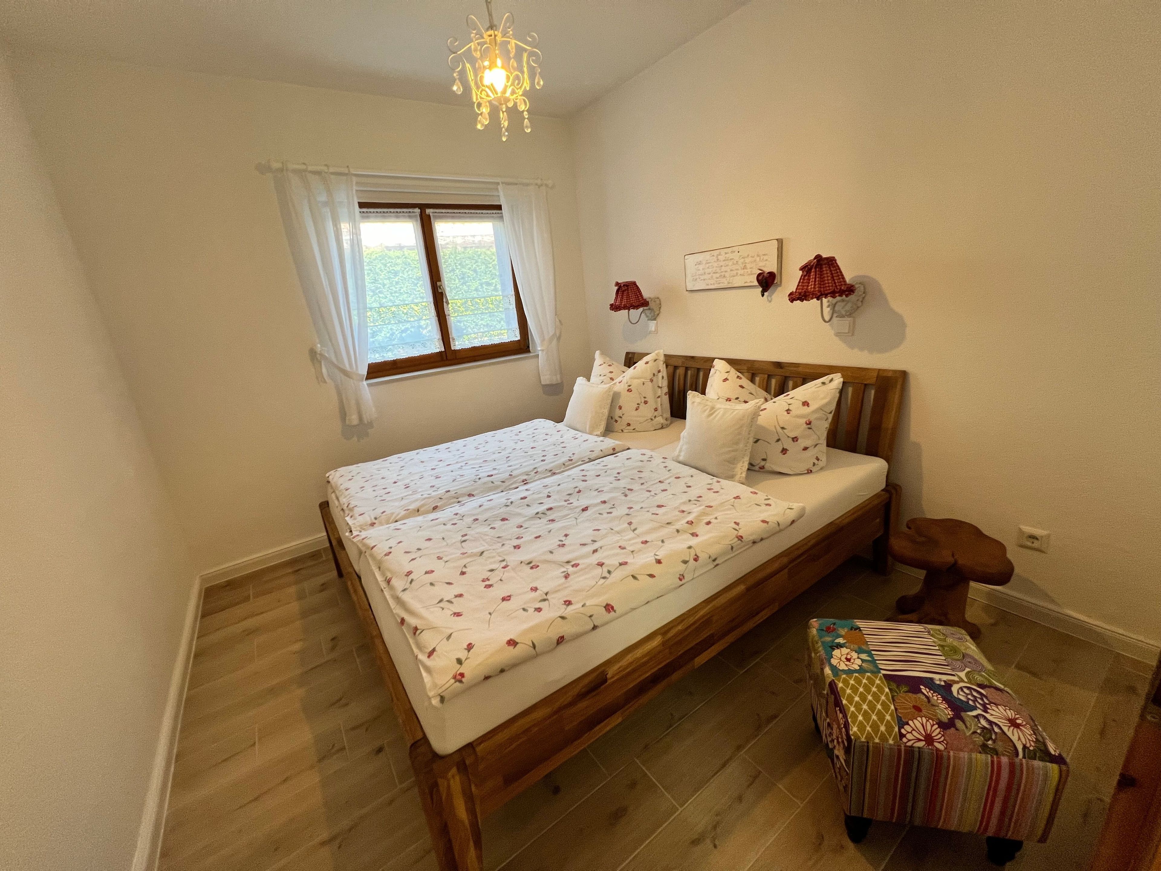 Ein Schlafzimmer mit einem Doppelbett, einem kleinen Nachttisch, einer Fensterbank, Vorhängen, einer Chandelieber und einer Wandleuchte.