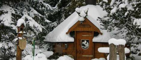 Una cabaña de madera con techo nevado, una puerta de madera con un escudo heráldico y un poste de madera con un letrero.