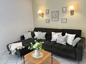 Un salon avec un canapé foncé, des coussins blancs et à motifs, une table basse en bois ornée d’un vase de fleurs et des lampes fixées au mur.