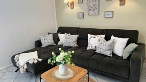 Un salon avec un canapé foncé, des coussins blancs et à motifs, une table basse en bois ornée d’un vase de fleurs et des lampes fixées au mur.