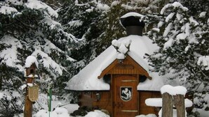 Une cabane en bois avec un toit enneigé, une porte en bois ornée d’un blason et un poteau en bois portant une enseigne.