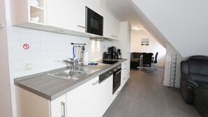 Una cocina moderna con armarios blancos, un lavamanos de acero inoxidable y un horno empotrado. Hay una zona de comedor con una silla negra y una mesa pequeña.