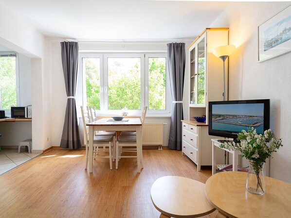 A well-lit room with a dining table, chairs, a television, and a view of greenery through the windows.