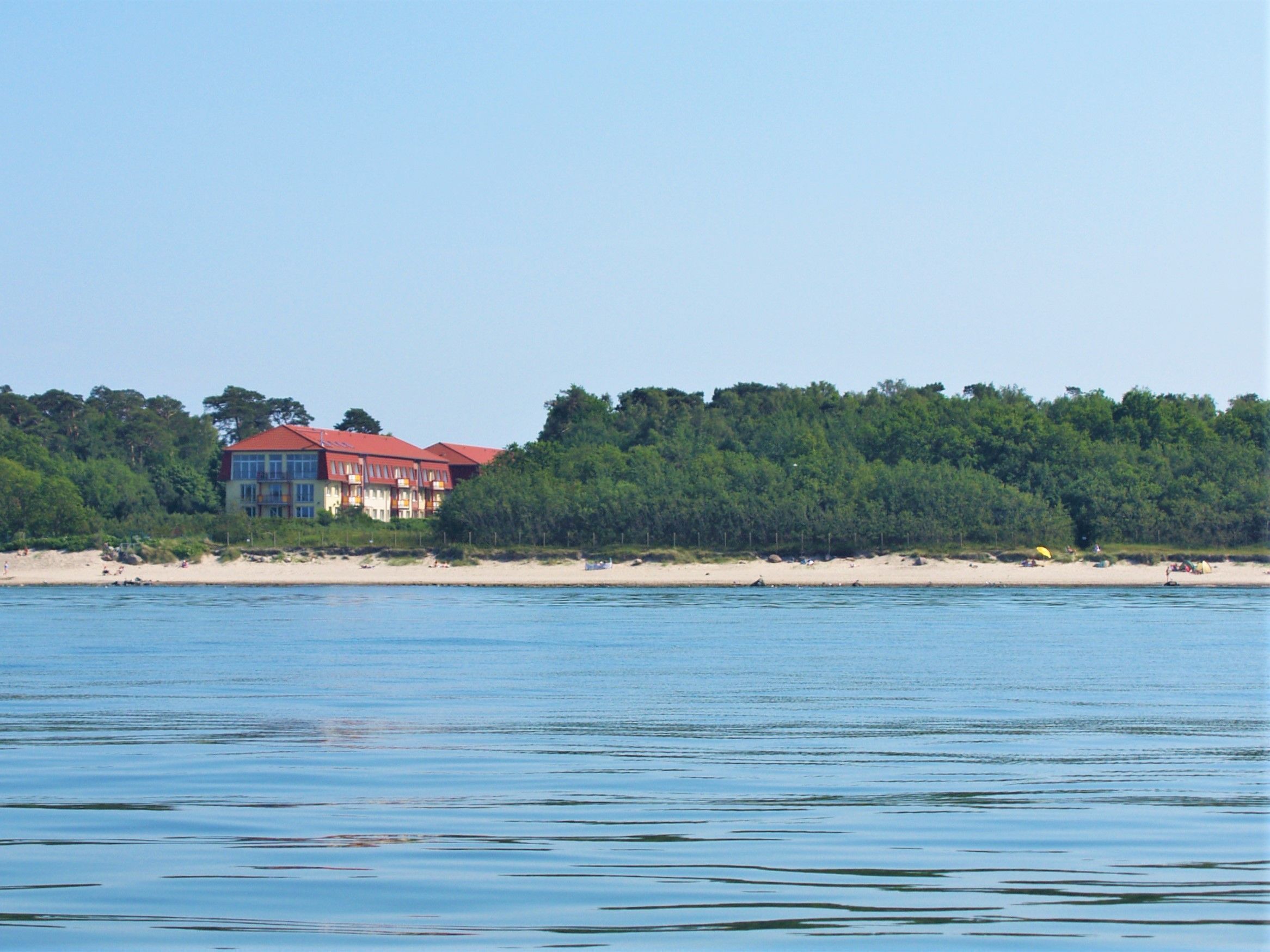 A large building with a red roof near a beach, surrounded by dense trees.