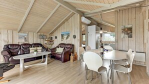 A cozy living room with a high ceiling, wooden walls, a leather sofa, a white table, and a dining area with chairs. - Lovely home in Rømø with kitchen (Rømø)