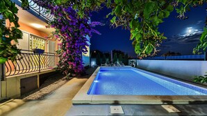Ein Schwimmbecken mit einer gefliesten Terrasse, umgeben von einem Garten mit violetten Blumen und einem Zaun.